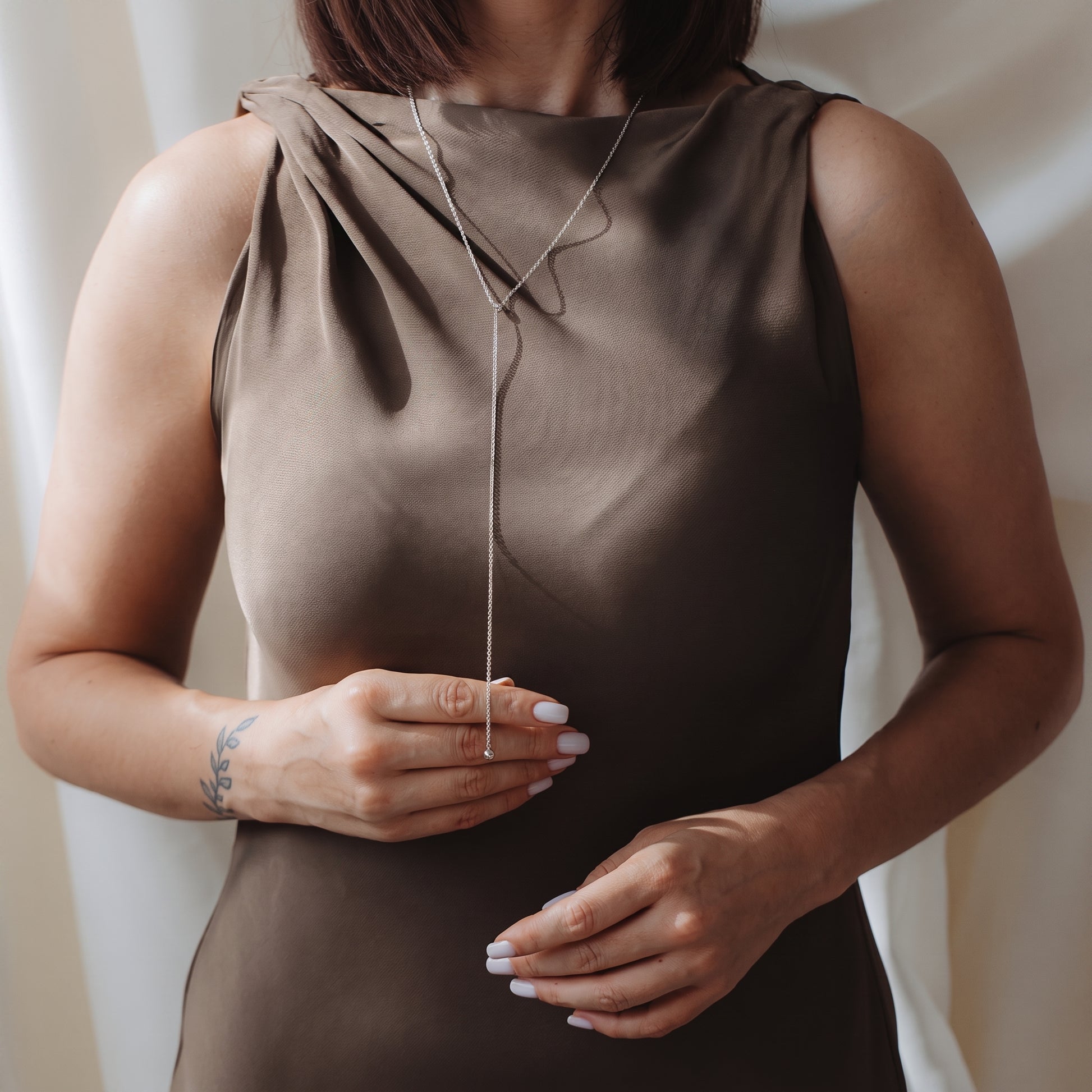 Zbliżenie modelki w eleganckim naszyjniku lariat bola ze srebra próby 925, założonym na sukienkę bez rękawów w kolorze taupe. Modelka delikatnie trzyma zwisający łańcuszek.