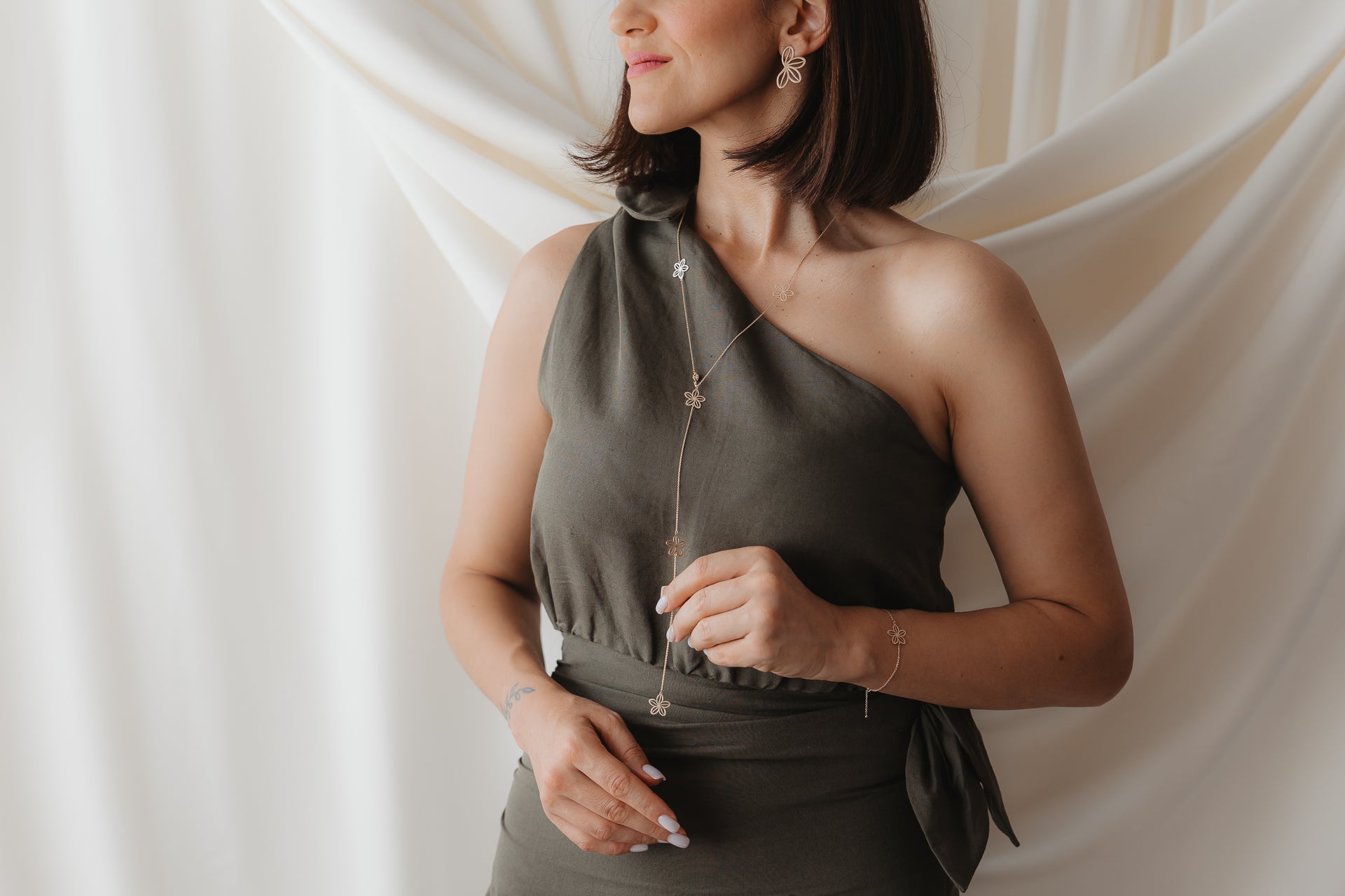 Zbliżenie na modelkę, która ma na sobie naszyjnik lariat Wildflower Maxi z żółtego złota 14K z delikatnymi zawieszkami w kształcie kwiatów, noszony na oliwkowej sukience na jedno ramię, na tle miękko udrapowanego kremowego tła.