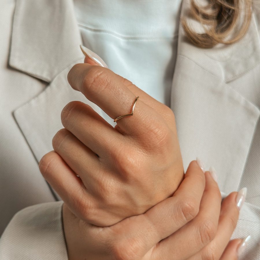 Zbliżenie na dłoń z prostym pierścionkiem w kształcie litery V, pozłacanym srebrem, na tle ubrania w neutralnych tonacjach.