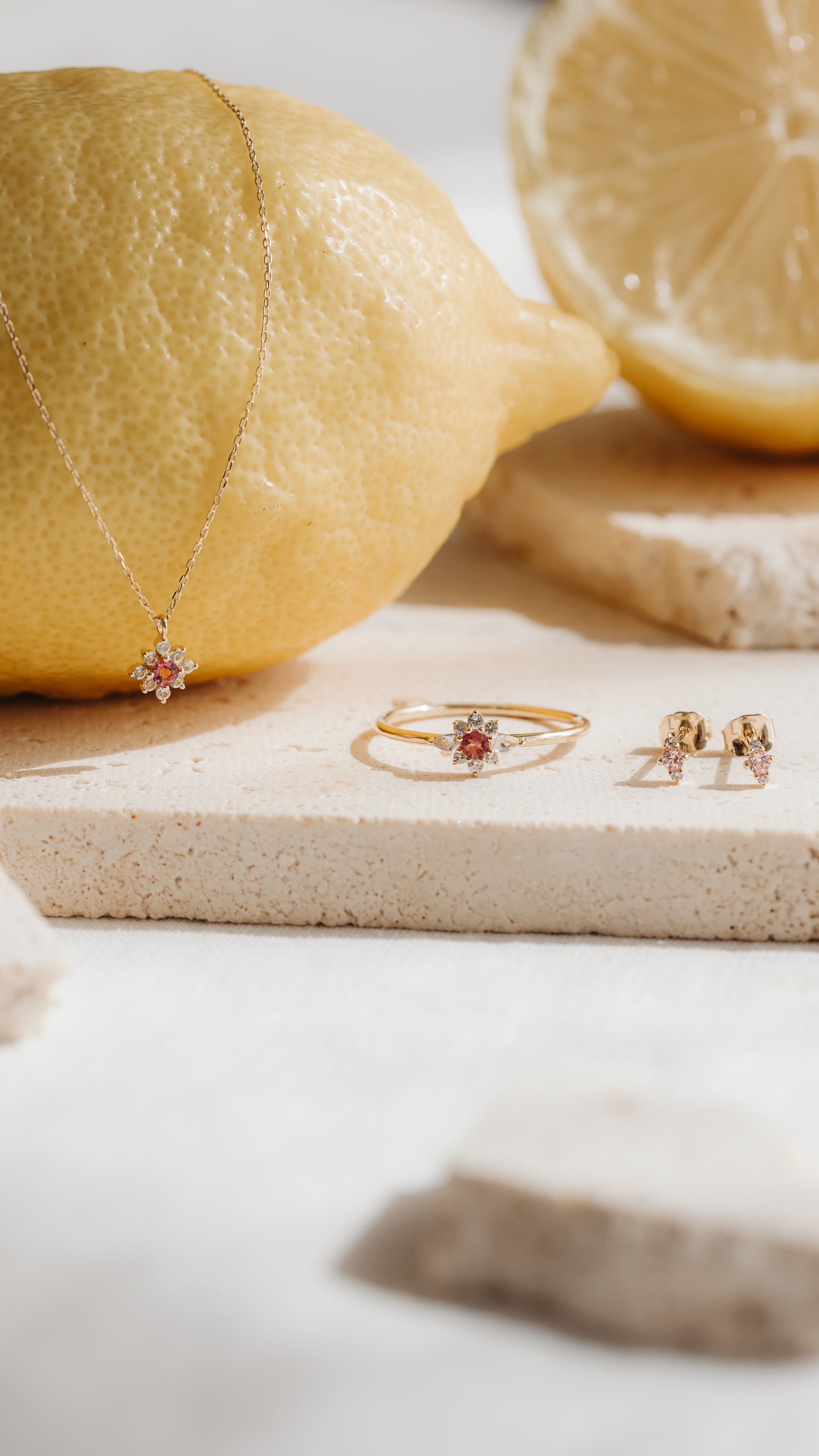 Kolczyki wkrętki Flora z żółtego złota 14K z różowym morganitem i białymi szafirami, zestylizowane obok pasującego pierścionka i wisiorka na kamiennych płytkach z cytrynami.
