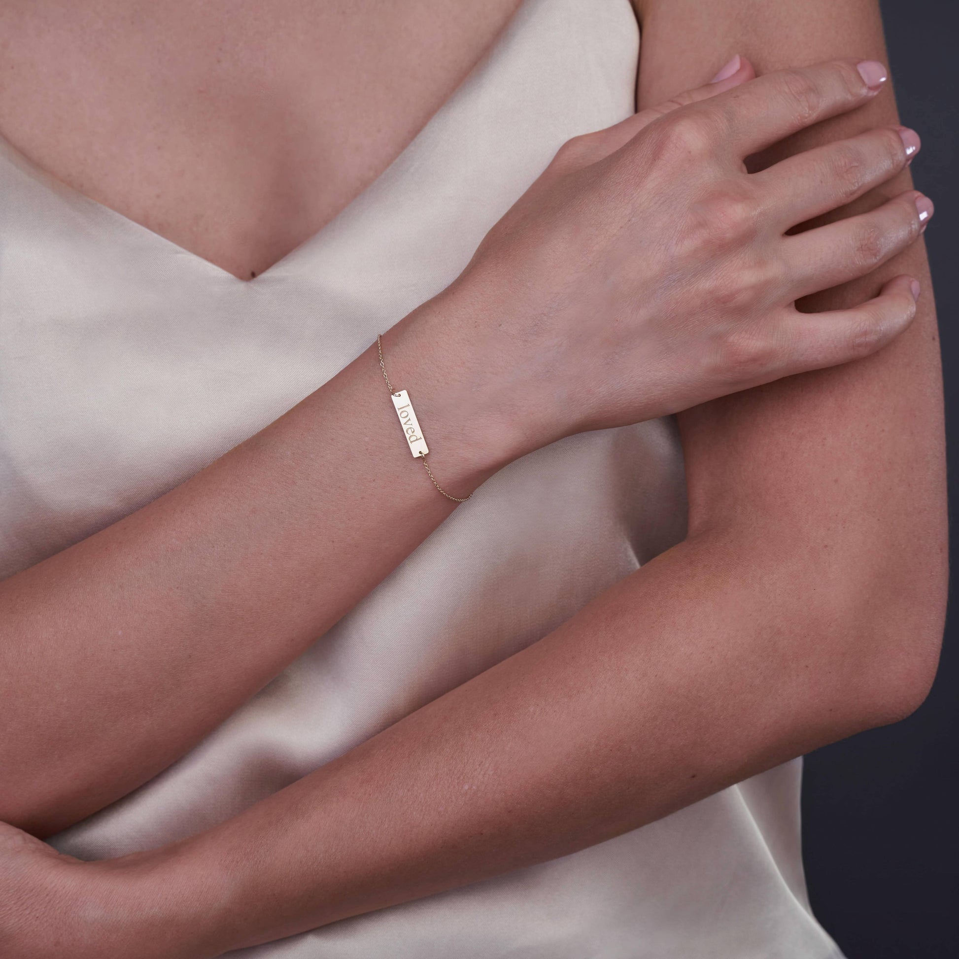 Zbliżenie osoby noszącej bransoletkę z 14K złota z wygrawerowanym słowem 'Loved', na tle neutralnym.