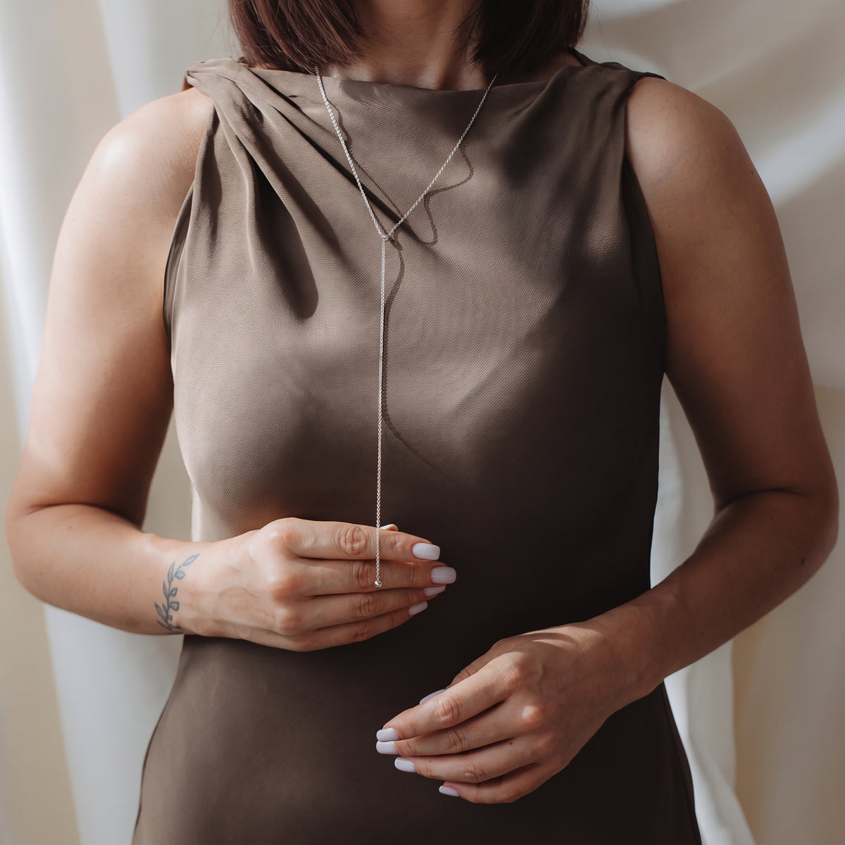 Zbliżenie modelki w eleganckim naszyjniku lariat bola ze srebra próby 925, założonym na sukienkę bez rękawów w kolorze taupe. Modelka delikatnie trzyma zwisający łańcuszek.