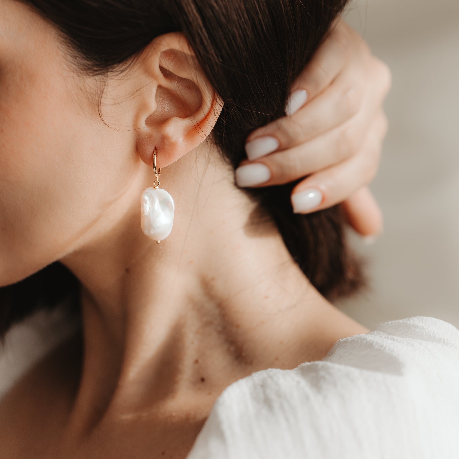 Zbliżenie ucha z pozłacanym kolczykiem-kołem ze srebra próby 925, ozdobionym dużą białą perłą barokową; dłoń z neutralnym manicure przy uchu w miękkim, naturalnym świetle.