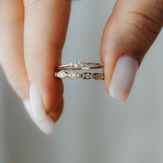 Zbliżenie dwóch diamentowych pierścionków do noszenia warstwowo z żółtego złota 14K, trzymanych między palcami — jeden z drobnym klastrem diamentów, drugi z motywami w kształcie markizy.